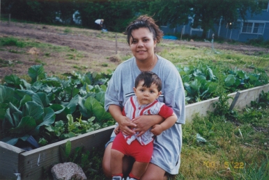 a Latino gardener kneels in front of her garden bed with her young son
