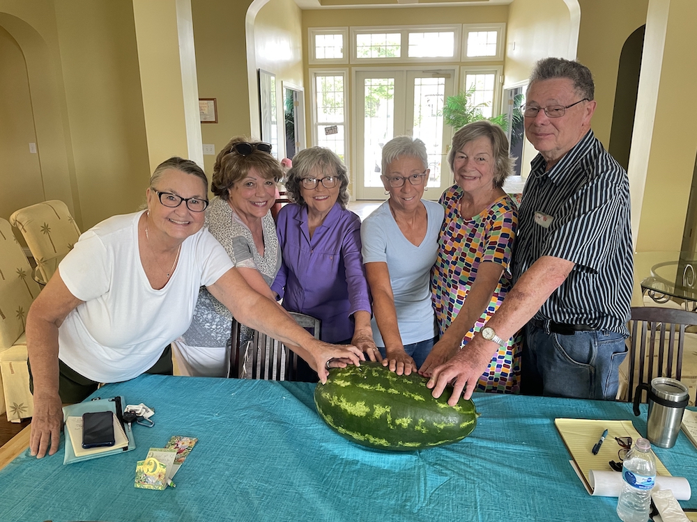 six older people stand in front of a table with their hands on a large watermelon